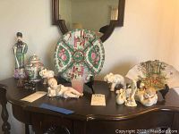 Wide shot of lot showing collector plate, figurines, lidded vase, and porcelain fan on table with mirror background