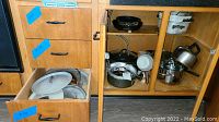 Cabinet and drawer opened showing various metal pots and pans, lids, and non-stick pans stored inside