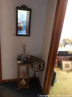 Wooden corner table with religious statuette, framed images, and other small decorative religious items positioned in a corner beneath a hanging mirror.