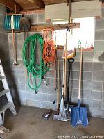 Photo of green garden hose and orange extension cord coiled and hung on a wooden rack on the wall with a ladder visible to the side. Various shovels and rakes standing on the floor below.