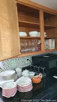 Kitchen cabinet and countertop showing stacked china plates and teacups, glass stemware, and a pumpkin-shaped creamer