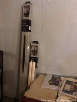 Two boxed Unique adjustable curtain rods with black finials leaning against the wall, along with beige and white folded linens on a table.