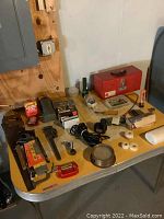 Yellow tabletop with red metal toolbox and assorted hand tools including pipe wrench, hand saw, drill bits, tape rolls, flashlight, and other hardware items.