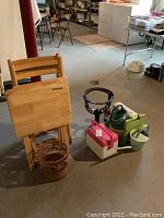 Photo showing wooden folding TV trays, folding chairs, wicker basket, plastic cooler, bocce ball set, watering cans, and metal table base arranged in basement.