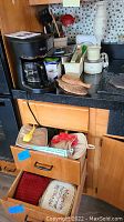 Photo shows a black Mr Coffee 5 cup coffeemaker, wooden kitchen utensils in a holder, some bottles and smaller kitchen items on countertop, with drawers open showing towels and pot holders.