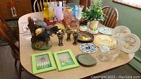 Overview of items on table including eagle decanter, wood figurines, glass vases, ceramic plates, faux plant, framed art, crochet pads