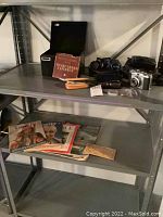 Shelving unit with various vintage items including cameras, binoculars, eyeglasses, reel films, and magazines on multiple shelves.