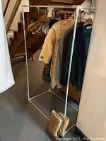 Side angle view of white metal rolling rack with several women's garments hanging and wool slippers on floor beneath.