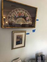 Framed hand painted decorative fan in gold-tone frame on wall above framed floral artwork