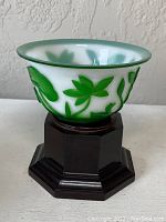 Side view of white frosted glass bowl with green floral relief design on octagonal dark wooden stand.