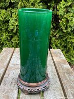 Full front view of tall cylindrical vase with green glaze on wooden stand.