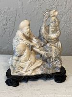 Front view showing three figures carved in stone with detailed facial expressions and clothing, sitting on a carved black wood stand.