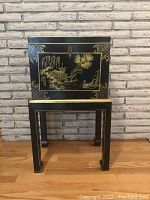 Front view of the black wooden storage cabinet with gold painted traditional Chinese scenes, sitting on matching stand.