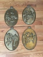 Four oval antique metal wall plaques arranged on wood floor, showing two pairs of different raised relief Chinese scenes.