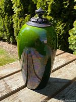 Green glazed oviform vase with carved wooden lid placed outdoors on a wooden table, showing the glossy finish and body shape.