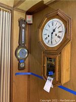View of both the regulator wall clock and wall barometer mounted on a wooden paneled wall, showing overall condition and scale.