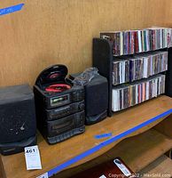 Compact stereo with open CD tray showing red CD, two speakers on either side, and wooden rack filled with CDs on right side.