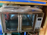 Oster French Door Countertop Oven with stainless steel body and glass double doors with handles. Oven interior includes racks and baking pan. Powers on with a digital control panel and multiple preset options.