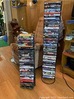 Image showing two tall black metal DVD racks fully loaded with various DVDs placed in a room with wooden and upholstered furniture.