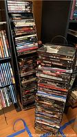 Stacked DVDs arranged on and around two tall vertical black metal DVD racks on a floor showing a variety of movie titles.