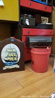 Photo showing wooden backgammon board with ship design labeled 'The Golden Clipper 1873', red bucket, and vintage radio on floor with other items around.