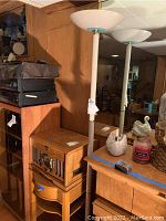 Photo showing two tall pole lamps with frosted glass shades placed next to a wooden cabinet with various decorative items on top.