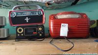 Three vintage styled radios placed side by side on wooden shelf. Ion Ford Mustang radio on top left, Studebaker cassette player below ion radio, and Magnavox red vintage radio/CD player on right.