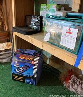Shelf setup showing Bissell Little Green Clean Machine rug cleaner in green plastic case with handle, Rolykit Deluxe Model paint roller system box below shelf, tool kit case black with screwdrivers and level visible, Pour N Paint roller paint system box on shelf, small white paint bucket on floor.