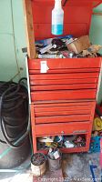 Red metal toolbox with open top compartment showing various hardware, wires, small boxed items, and loose tools inside.