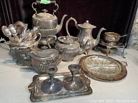 Shows a broad view of the lot with kettle, teapots, silverware holder, trays, candlesticks, and glass dish.