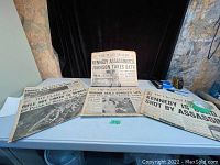 Five newspapers laid out on a table, with visible headlines about Kennedy's assassination and related events, showing the overall lot condition and content.