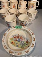 Photo showing 14 Bunnykins cups and 1 Bunnykins bowl with center scene and rabbit border.