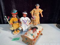 Four indigenous dolls: three standing in traditional clothing, one lying in a decorated wooden crib with fur and feathers