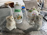 Photo of four tea and coffee pots on glass table, including two ceramic cat-shaped pots and two antique porcelain pots with floral decoration.