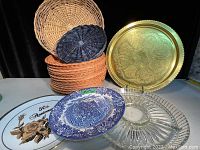 Photo showing 12 wicker plate holders, blue and white ceramic plate, clear crystal divided dish, gold-toned metal tray, and additional woven baskets grouped together.