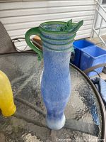View of the tall light blue blown glass vase with green handle and rim accent sitting on a glass table outdoors.