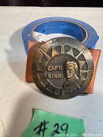 Front view of metal belt buckle showing raised Capt. Kirk portrait and text, leather backing visible underneath.