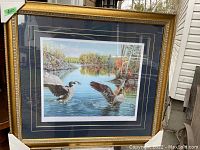 Full view of the gilded wood frame containing the print of geese over a river scene with trees in autumn colors.