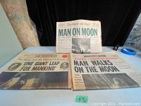 Photo showing three newspapers from 1969 reporting the Apollo 11 Moon landing with visible headlines and typical age wear.