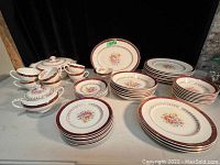 Partial antique china dish set displayed with plates stacked, cups and bowls grouped closely, serving tureens with lids visible. Items show floral designs with maroon and gold borders.