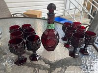 Photo showing ruby red decanter and 12 matching glasses arranged on a glass table outdoors
