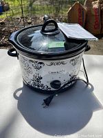Crock Pot slow cooker with glass lid and instruction manual placed outside on a white table in daylight.