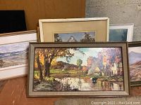 Photo showing a collection of framed landscape artworks including a river and cottages scene in autumn colors and additional prints behind it.