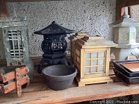 Photo showing black iron pagoda lantern, tan wooden lantern, white ceramic lantern, and assorted pots and planters on shelf.