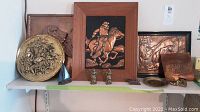 Shelf display showing multiple brass and copper decorative items including plaques, figurines of ships, soldiers, and small train engine.