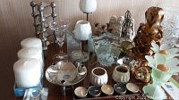 Wide view of the assorted candle holders and candles on a wooden surface showing various materials and styles including ceramic, glass, metal, and decorative figurines.