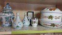 Shelf showing two oil and vinegar cruets with floral design, blue and white lidded jar, and white ceramic container with metal lid and fruit decoration.