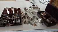 Wide view of two open presentation boxes of spoons, loose souvenir spoons, serving utensils, and creamer set on table