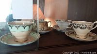 Front view of 5 assorted teacups and saucers with gold accents displayed in cabinet