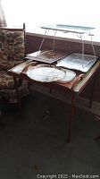 Photo showing wooden folding card table holding several trays: painted street scene tray elevated on metal stand, the pewter tray, and wooden tray.